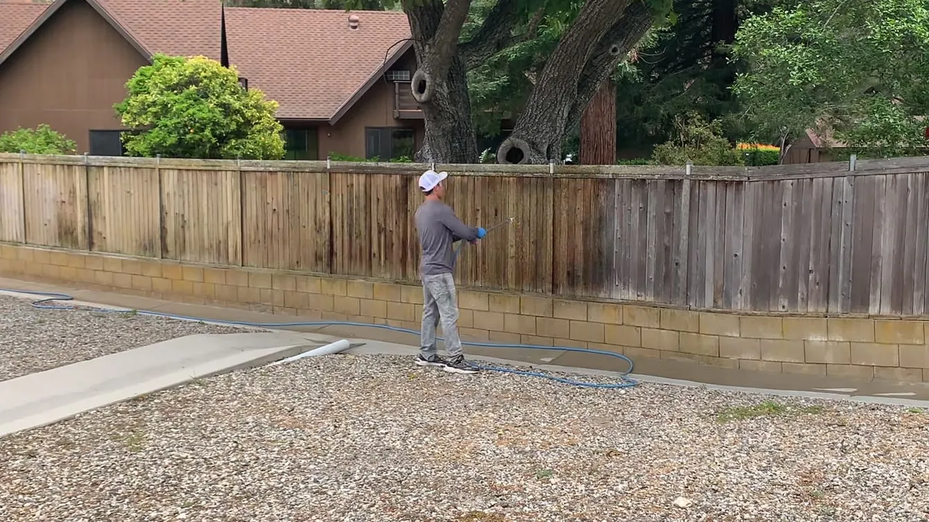 A worker pressure-washing a weathered wood fence in Santa Barbara, the cleaned wood meeting the gray weathered boards in a sharp line