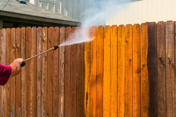 A pressure washer stripping years of gray weathering off a wood fence, revealing rich, warm clean wood behind the spray