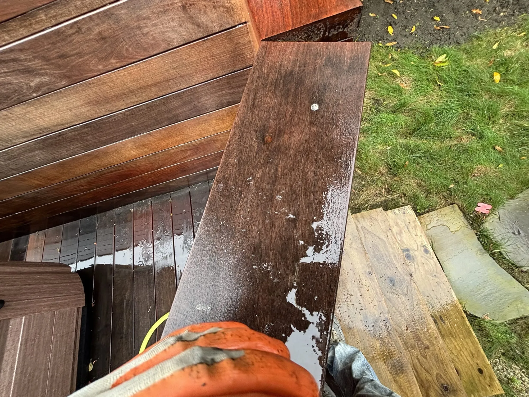 A gloved hand wiping fresh oil-based stain onto a deck board, the wood turning a wet, rich reddish-brown